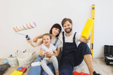 Cheerful caucasian family of father, mother and cute little girl daughter sitting on floor in white light room background with paint rollers in their hands.