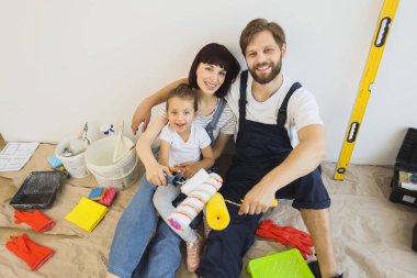 Happy family of father, mother and cute little daughter playing while enjoying renovating in their bright room, putting their hands together with paint rollers.