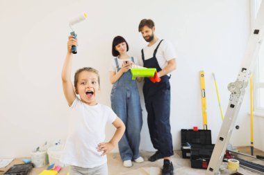Happy overjoyed caucasian small girl raising her hand with paint roller while blurred mom and daddy standing on the background and looking for interior design idea for renovation on phone.