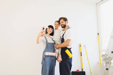 Lovely happy caucasian family of young mother, bearded father in overalls and their little daughter making selfie on smartphone, relaxing after painting wall with paint roller in white