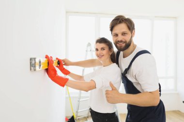 Repair home concept. Young caucasian couple are measuring a wall with measuring tape. Bearded smiling man showing thumb up. Activities after relocation and renovation.