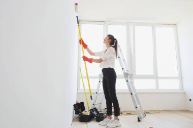 Beautiful young caucasian happy woman makes repairs in apartment while painting interior wall with paint roller in new house. Home decoration concept.