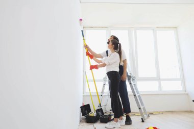 Side view of young caucasian couple, beautiful woman and bearded man in overalls with paint rollers painting wall near ladder in white colour.