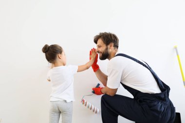 Happy family, young caucasian father and his cute child daughter standing and paints the wall with white paint giving high five. Repair in the apartment concept.