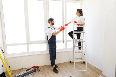 Attractive young couple in rubber gloves with detergent and rag washing windows together. Beautiful woman in casual clothes and bearded man in overalls cleaning their house after repair.