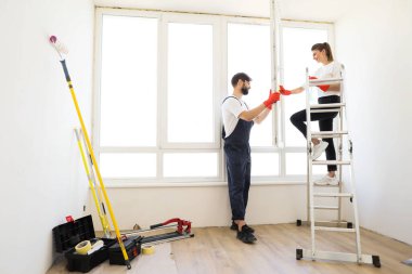 Attractive young couple in rubber gloves with detergent and rag washing windows together. Beautiful woman in casual clothes and bearded man in overalls cleaning their house after repair.