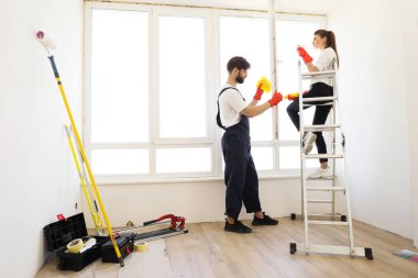 Young family of two, pretty woman sitting on ladder and bearded man household chores, they wash the window in their house along with cleaning agent and yellow house cleaning rags.