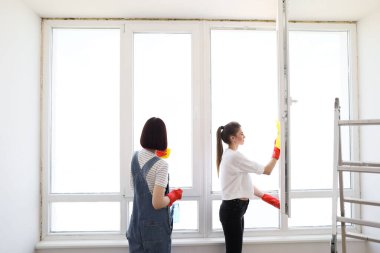 Hard working girls using spray. Young females washing window while working together. Young caucasian women, sisters or friends cleaning window together in room.