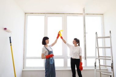 Beautiful young women in rubber gloves with detergent and rag washing windows together giving high five. Attractive caucasian girls making house cleaning after repair.