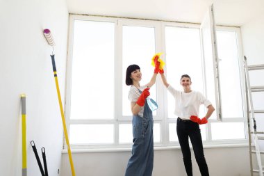 Young females washing window while working together giving high five. Young caucasian women, sisters or friends cleaning window together in room. Hard working girls using spray.
