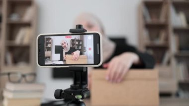 Focus on phone screen , muslim woman recording video on phone camera while unpacking box with new wireless laptop. Female influencer sharing with subscribers her positive feedback about new order.