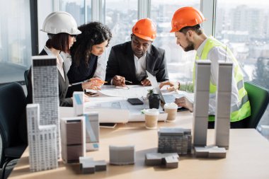 Group of four multiracial people architects sitting at desk with lots of blueprints and architecture design of buildings with residential project maquette at open space office with panoramic windows.