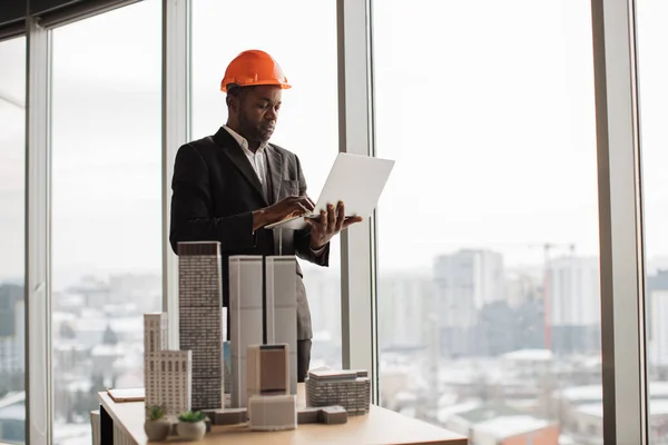 Handsome chief architect african man in business suit and protective helmet with laptop pc working with city buildings model as urban planner at office interior with panoramic city view.