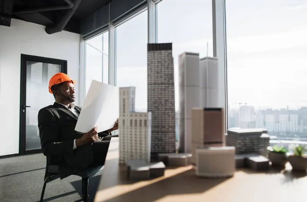 Office interior with panoramic city view. Handsome chief architect man in business suit and protective helmet holding blueprint sitting at desk working with city buildings model.
