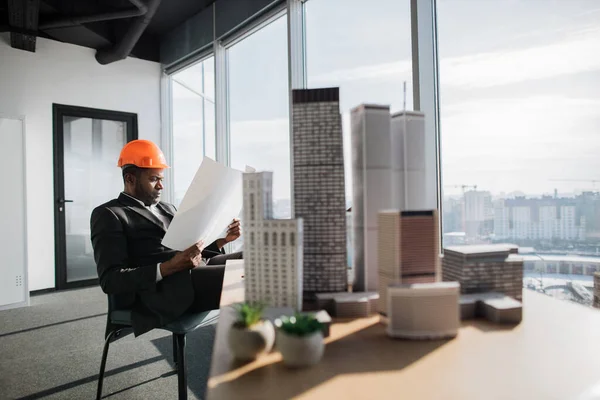 Office interior with panoramic city view. Handsome chief architect man in business suit and protective helmet holding blueprint sitting at desk working with city buildings model.