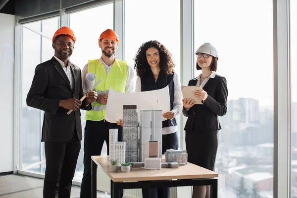 Happy multiracial architects, engineers and designers standing near skyscrapers buildings maquette during meeting at boardroom. Men and women colleagues leading meeting for architectural project.