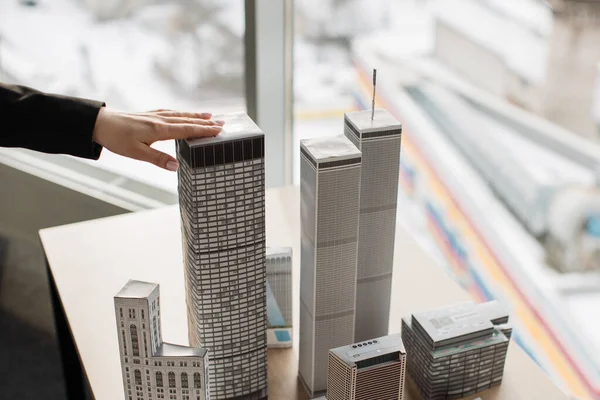 Architectural bureau office with panoramic windows and blurry cityscape. Hand of young woman touching one of building complex prototype project of residential or business district, city model.