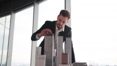 Confident businessman real estate agents in black stylish formal suit standing while touching 3d model skyscraper of city architecture design at office with panoramic city view.