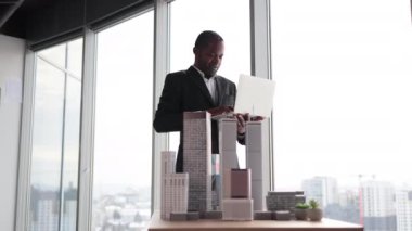 Smiling african american business man real estate agents in black stylish formal suit standing with laptopnear 3d model skyscrapers of city architecture design at office with panoramic city view.