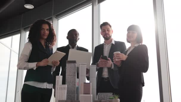 由四个不同种族的商人组成的团队在办公室的会议室开会 全景窗模糊 城市景观朦胧 他们站在桌旁 摩天大楼的礼仪展示着大拇指 — 图库视频影像