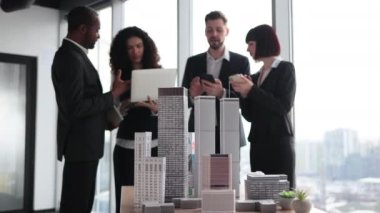 Close up of residential project maquette. Beautiful confident curly business lady showing her financial and sales report on laptop pc for her focused high-skilled multiracial business colleagues.
