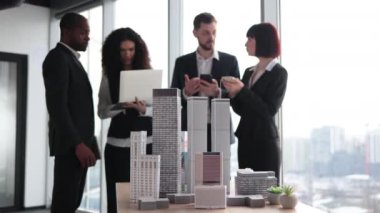 Close up of residential project maquette. Beautiful confident curly business lady showing her financial and sales report on laptop pc for her focused high-skilled multiracial business colleagues.