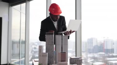 Focused african man in black suit and orange hardhat using digital laptop looking at design sustainable 3D megalopolis model on desk at open space office with panoramic windows with blurry cityscape