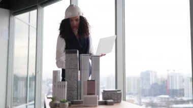 Focused woman in formal wear and white hardhat using digital laptop looking at design sustainable 3D megalopolis model on desk at open space office with panoramic windows with blurry cityscape.