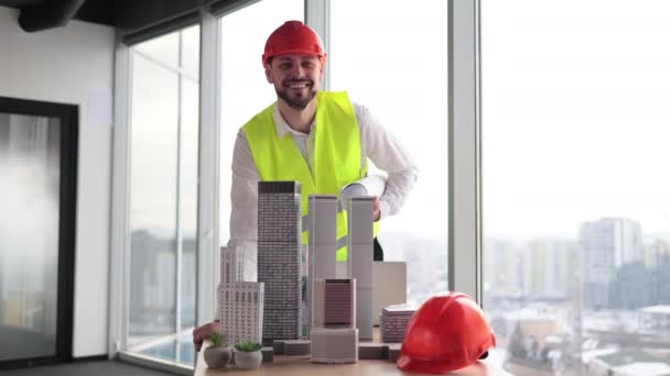 在具有全景城市视野的现代建筑办公室中 合格的建筑师或工程师身穿反光背心 头戴硬礼帽检查摩天大楼模型 有胡子的有吸引力的男人控制工作过程 — 图库视频影像