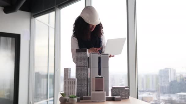 ビジネススーツの魅力的な若い女性建築家とパノラマビューとオフィスインテリアで都市プランナーとして都市の建物モデルと作業ノートパソコンのPcと白の保護ヘルメット — ストック動画