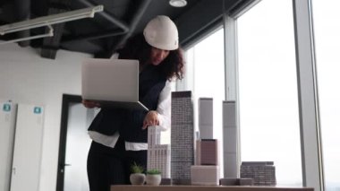 Genç bayan mimar mimari bürosunda panoramik şehir görüşü olan modern mimari ofiste gökdelen modelini kontrol eden dizüstü bilgisayar kullanıyor. Takım elbiseli ve kasklı çekici bir kadın..