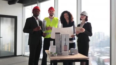 Group of four colleagues standing near building complex prototype project during successful brainstorming session at architectural bureau office. Construction team standing with blueprints and gadgets