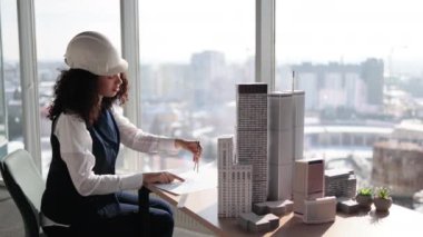 Curly female engineer working on building complex prototype project of residential or business district, measuring blueprint city model using compass at office with panoramic windows