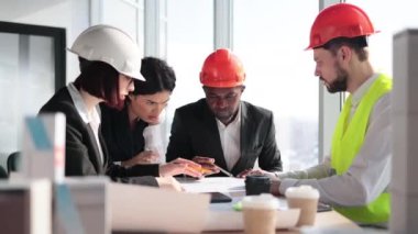Group of four multiracial people architects sitting at desk with lots of blueprints and architecture design of buildings with residential project maquette at open space office with panoramic windows.