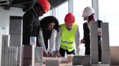 Çok ırklı inşaat işçileri ve mimarlar ellerinde bina projelerinin planları, cihazları ve tasarımlarıyla masanın yanında duruyorlar. İnşaat projesinde çalışan iş adamları