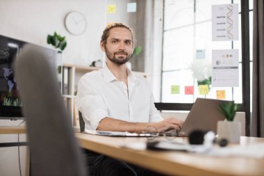 Modern ofiste poz veren şık resmi gömlekli bir iş adamı. Yakışıklı, genç, gülümseyen sakallı iş adamı. Masasında kağıtlar ve modern bilgisayarla oturuyor..