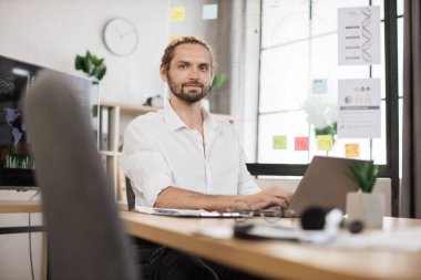 Business guy in stylish formal shirt posing at own modern office. Handsome young smiling bearded businessman sitting at desk with papers and modern laptop.