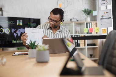 African american male office worker in eyeglasses striped shirt and headset talking and showing financial report during video conference on modern laptop. Online communication concept.