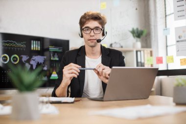 Young businessman in headset using laptop computer during video call working in office. Concentrated adult successful man wearing official suit sitting at wooden desk indoor holding pen in hands.