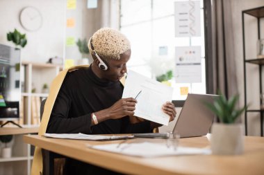 Serious marketing african man in suit and headset talking to client on video call, presenting paper graphic reports, showing, explaining diagram. Employee, manager attending online meeting