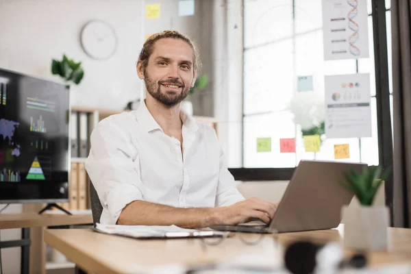 stock image Attractive positive caucasian guy in white shirt posing at own modern office. Young bearded businessman with shining smile, sitting at desk with papers and modern laptop.