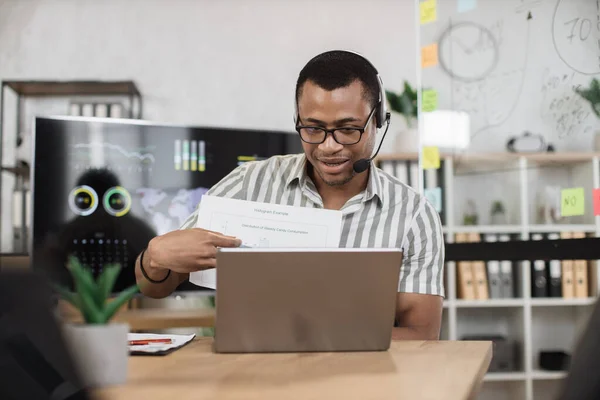 African american male office worker in eyeglasses striped shirt and headset talking and showing financial report during video conference on modern laptop. Online communication concept.