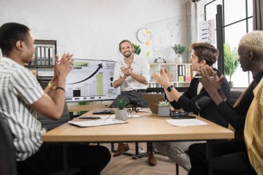 Multi ethnic male partners in formal clothes applauding together while sitting at office. Competent financiers businessmen with modern gadgets on table celebrating successful completion of project.