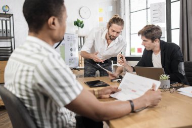 Group of competent male traders sitting together at office desk with various graphs and charts and working on gadgets. Multiracial partners during working process indoors.