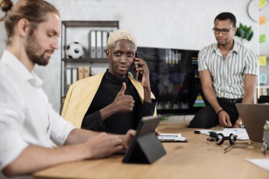 Focused african man speaks on the phone while his male colleagues listening speech of african american businessman. Company managers working at office room showing thumb up. Deadlines concept.