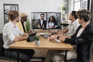 Konferans odasında odaklanmış çok ırklı iş adamları, ekrana bakıyorlar, başka bir ekiple görüntülü görüşme yapıyorlar, Afrikalı kadın ve beyaz adam, iletişim kurmak için akıllı video teknolojisi kullanıyorlar..