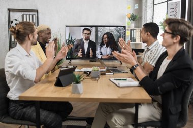 Group of four skillful multiethnical businesspeople clapping hands and looking at screen, having video conference in boardroom, with their colleagues explaining some financial charts.