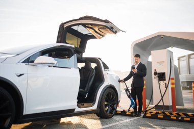 İyi giyimli erkek girişimci modern akıllı telefondan bataryayı kontrol ederken açık hava istasyonunda lüks elektrikli araba şarj ediyor. Alternatif otomobil ve ekoloji koruma kavramı.