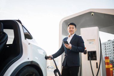 İyi giyimli erkek girişimci modern akıllı telefondan bataryayı kontrol ederken açık hava istasyonunda lüks elektrikli araba şarj ediyor. Alternatif otomobil ve ekoloji koruma kavramı.