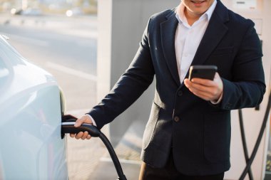 Siyah takım elbiseli mutlu Asyalı adamın beyaz lüks arabayı şarj etmesi ve modern akıllı telefon kullanması. Elektrikli araç teknoloji şarj etme kavramı.
